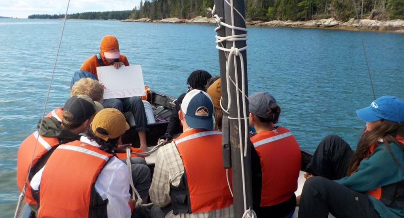 A group of people on a sailboat wear life jackets and crowd around an someone holding a whiteboard giving a lesson. 